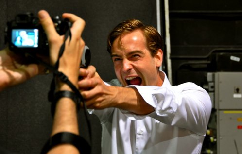 Behind the scenes on Slingers, Tom as Frank is wearing a white shirt, pointing a gun, and yelling