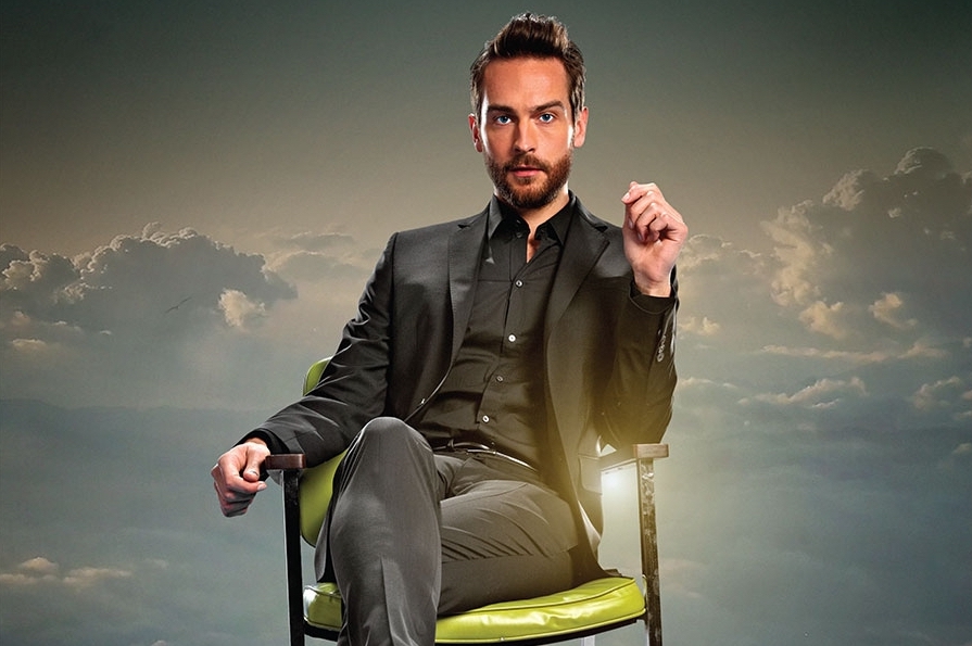 Tom sits in a lime green chair, in a killer black suit, with clouds in the background